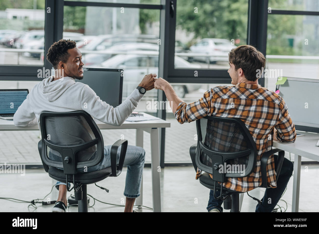 young multicultural programmers doing fist bump while sitting in office together Stock Photo
