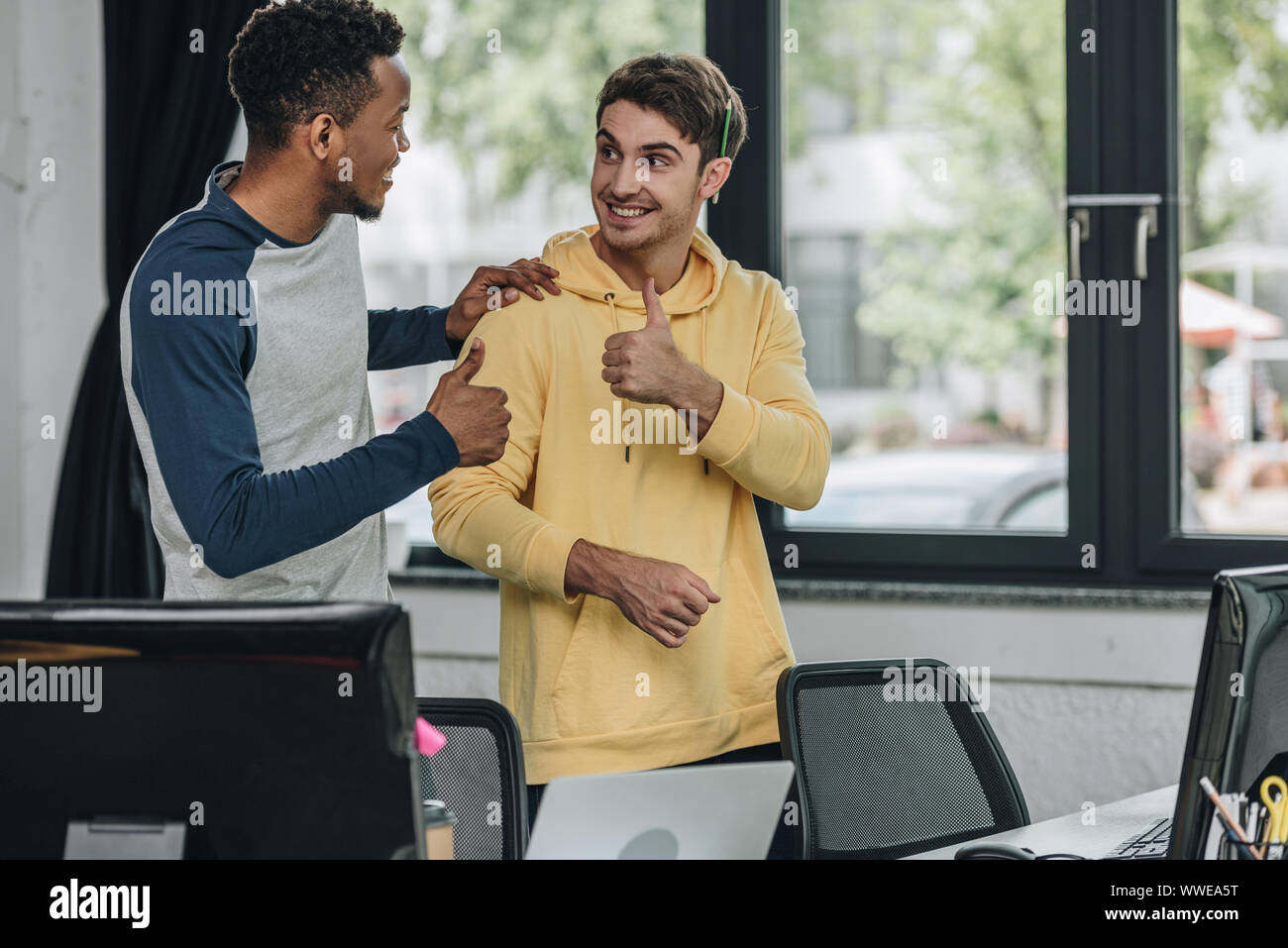 cheerful multicultural programmers looking at each other and showing thumbs up Stock Photo