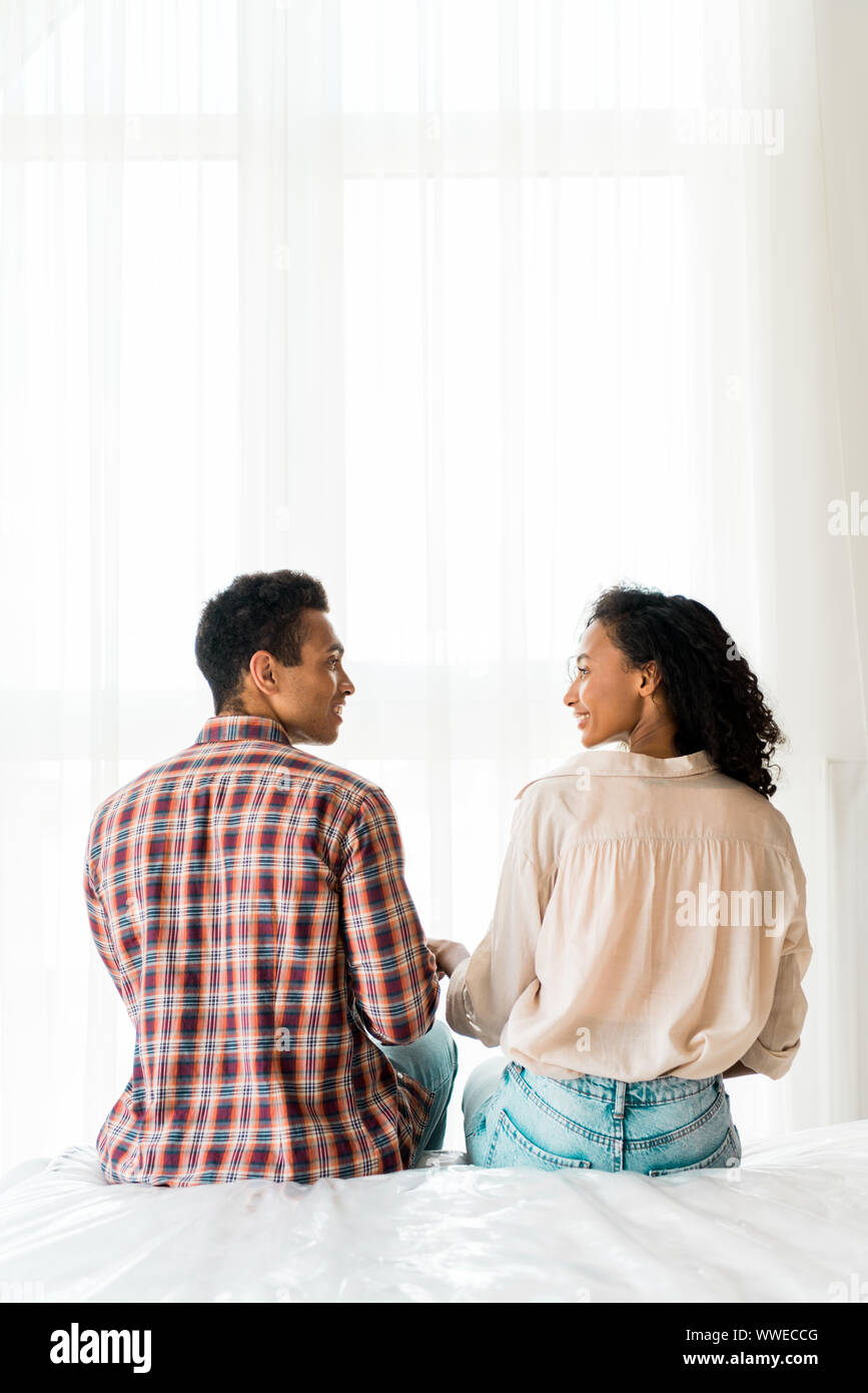 african american husband and wife sitting on bed, holding hands and ...