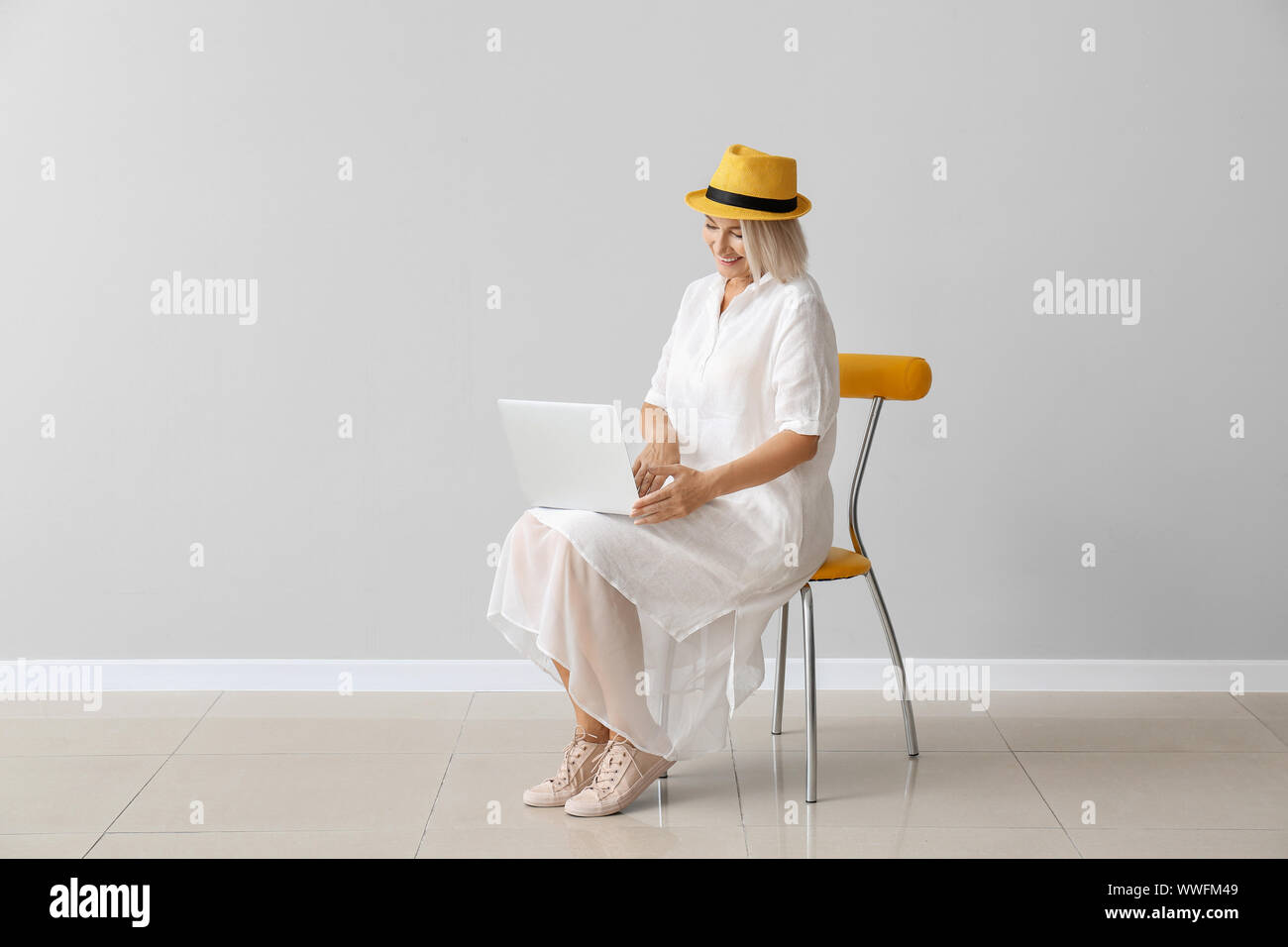 Mature woman with laptop sitting on chair against light wall Stock ...
