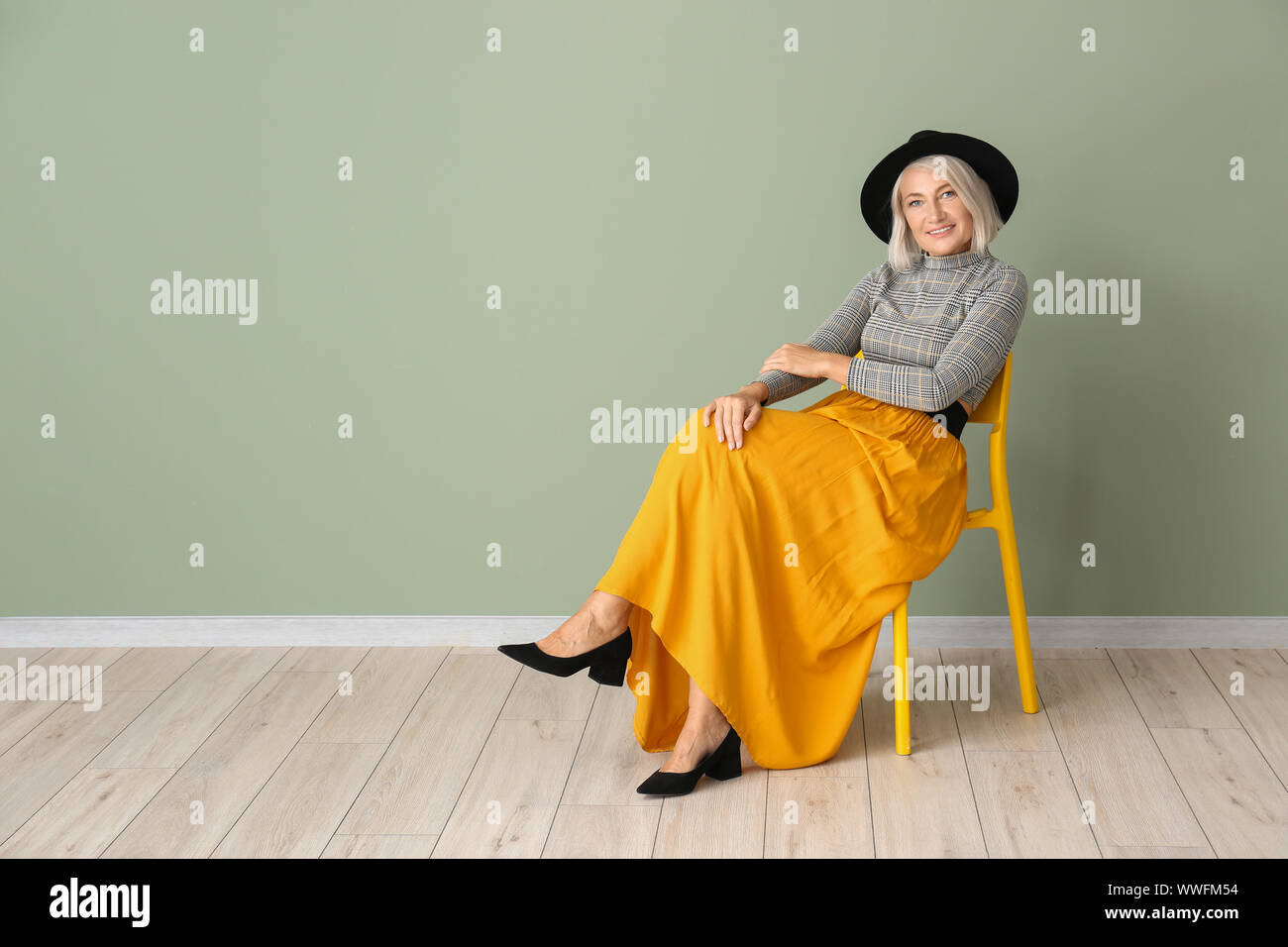 Stylish mature woman sitting on chair against color wall Stock Photo ...