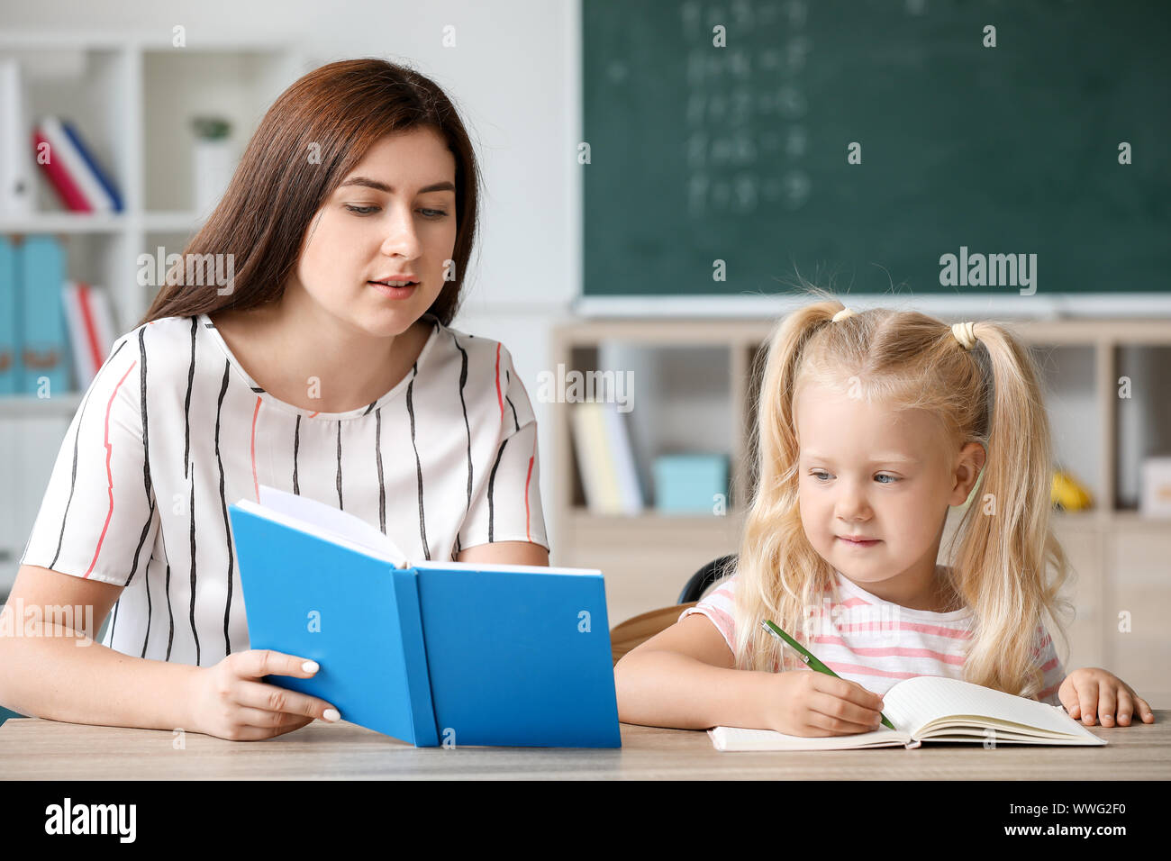 Teacher helping little girl to do task in classroom Stock Photo - Alamy