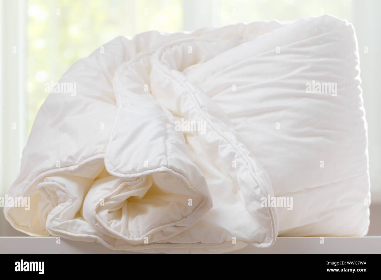 A duvet lies on a chest of drawers against a blurred window. Household. Stock Photo