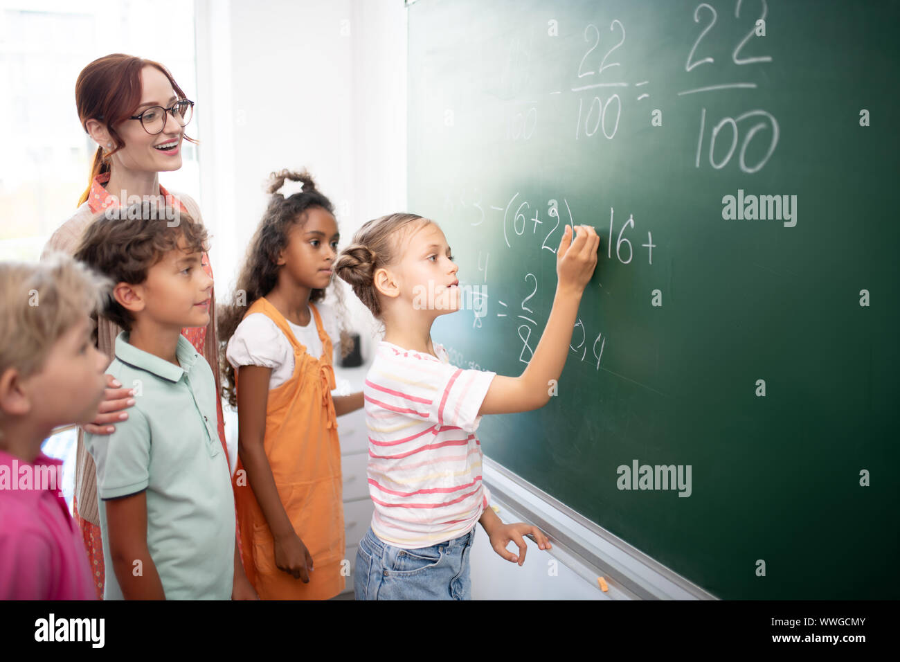 Smart girl writing down numbers on blackboard during math class Stock Photo