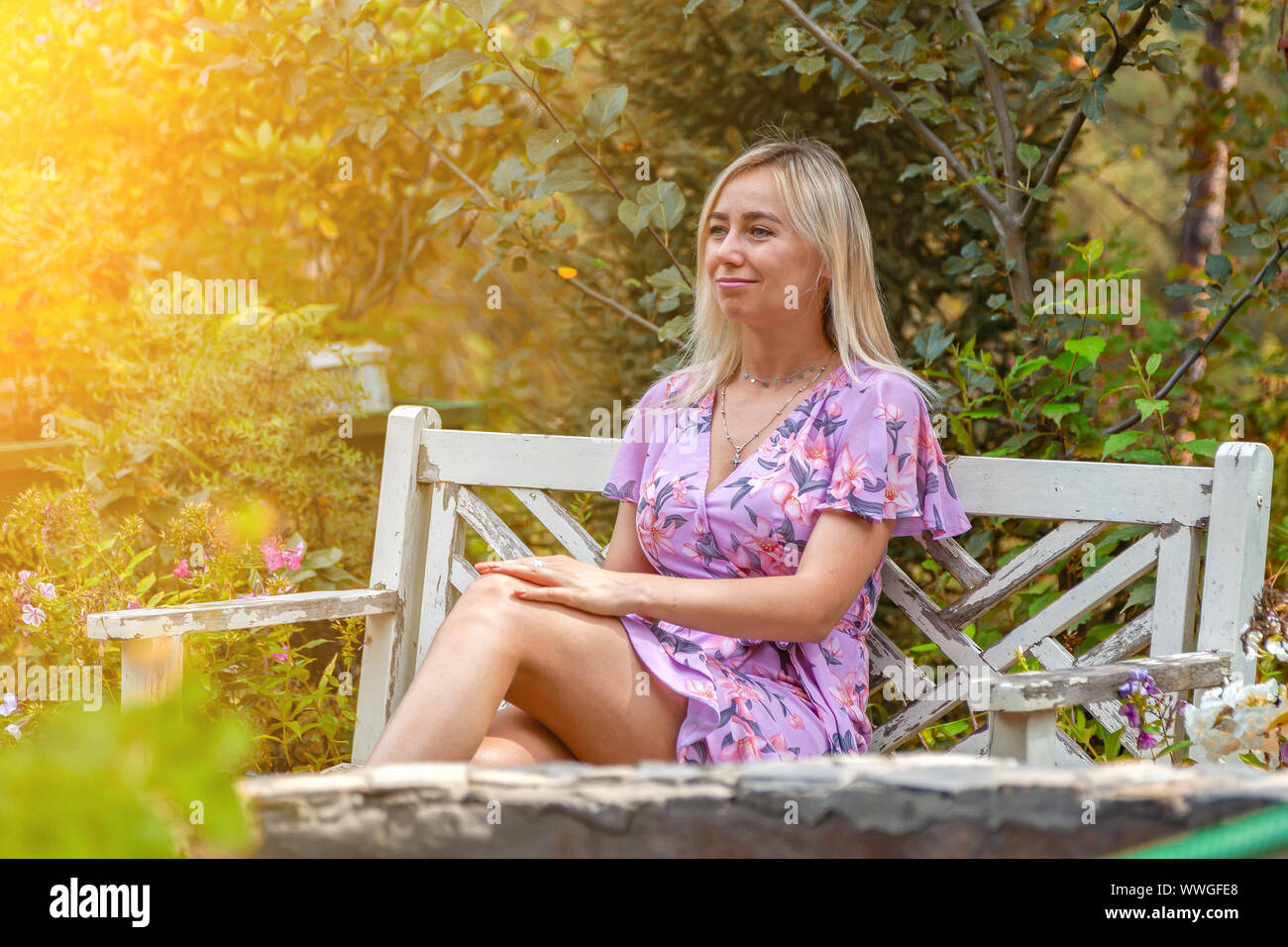 A young blonde girl in a pink dress sits on a white wooden bench in the ...