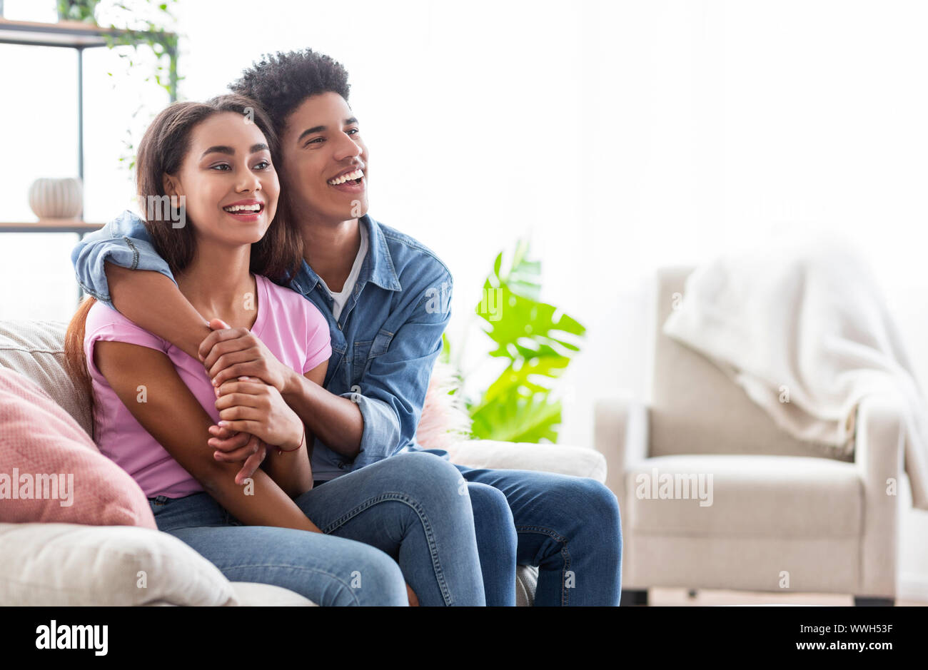 Black cheerful teens sitting on couch, cuddling and looking aside Stock Photo