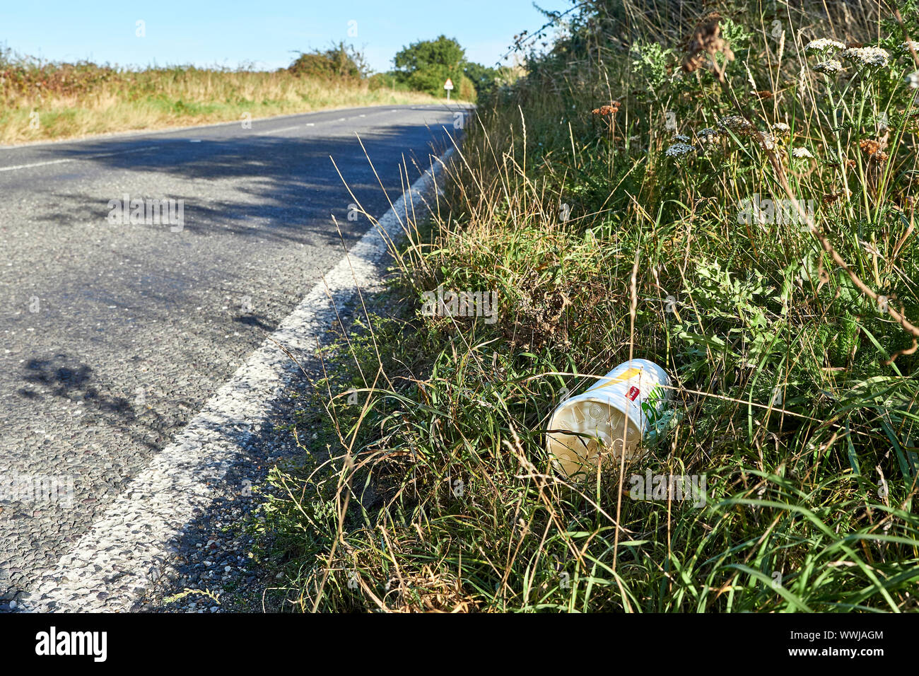 A low angled view of a cardboard cup that has been thrown out from a vehicle lead in the grass on the side of a main road that has no traffic on it Stock Photo