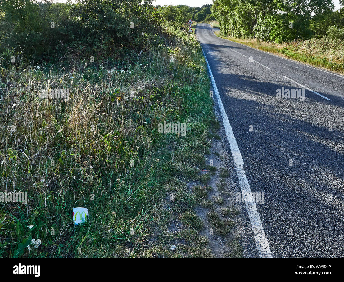 A cardboard cup that has been thrown out from a vehicle lead in the grass on the side of a main road that has no traffic on it Stock Photo