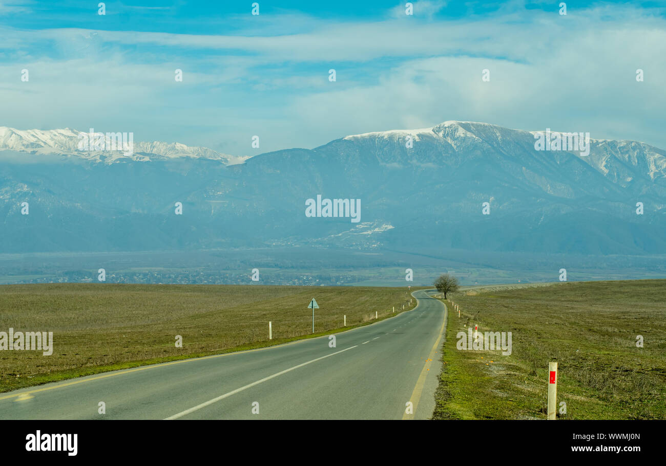 Mountain village with snowy peaks in the background long asphalt road in foreground. Stock Photo
