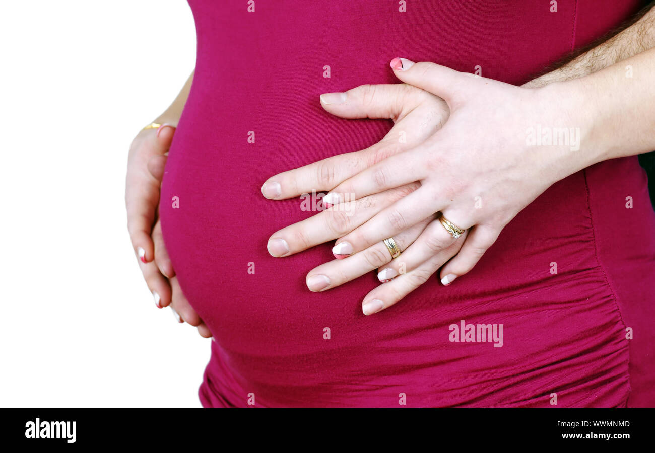 Beautiful studio shot of a 7 months pregnant married woman's belly in pink shirt with her hands and his hands intertwined, rings on both. Stock Photo