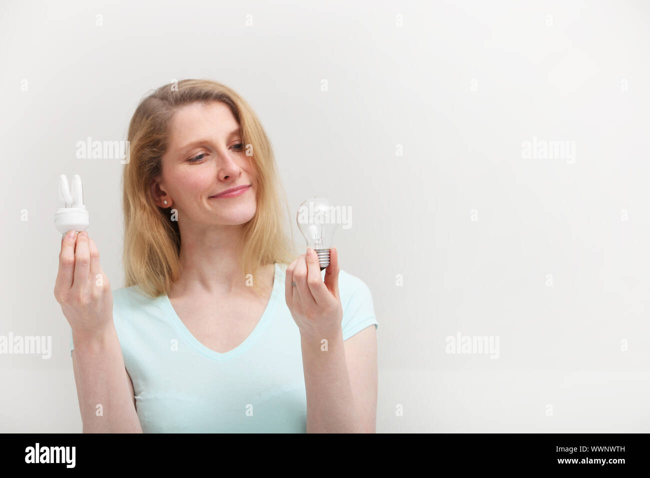 Woman holding an eco-friendly spiral fluorescent bulb in one hand looks at an old inefficient incandescent bulb in her other hand with regret as she m Stock Photo