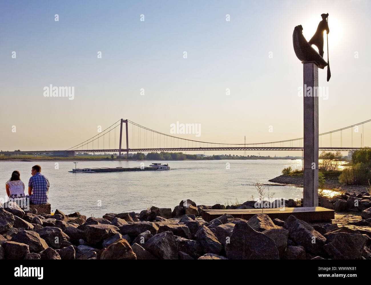 two young people sitting on the Rhine river bank next to sculpture ferryman, Emmerich, Germany Stock Photo