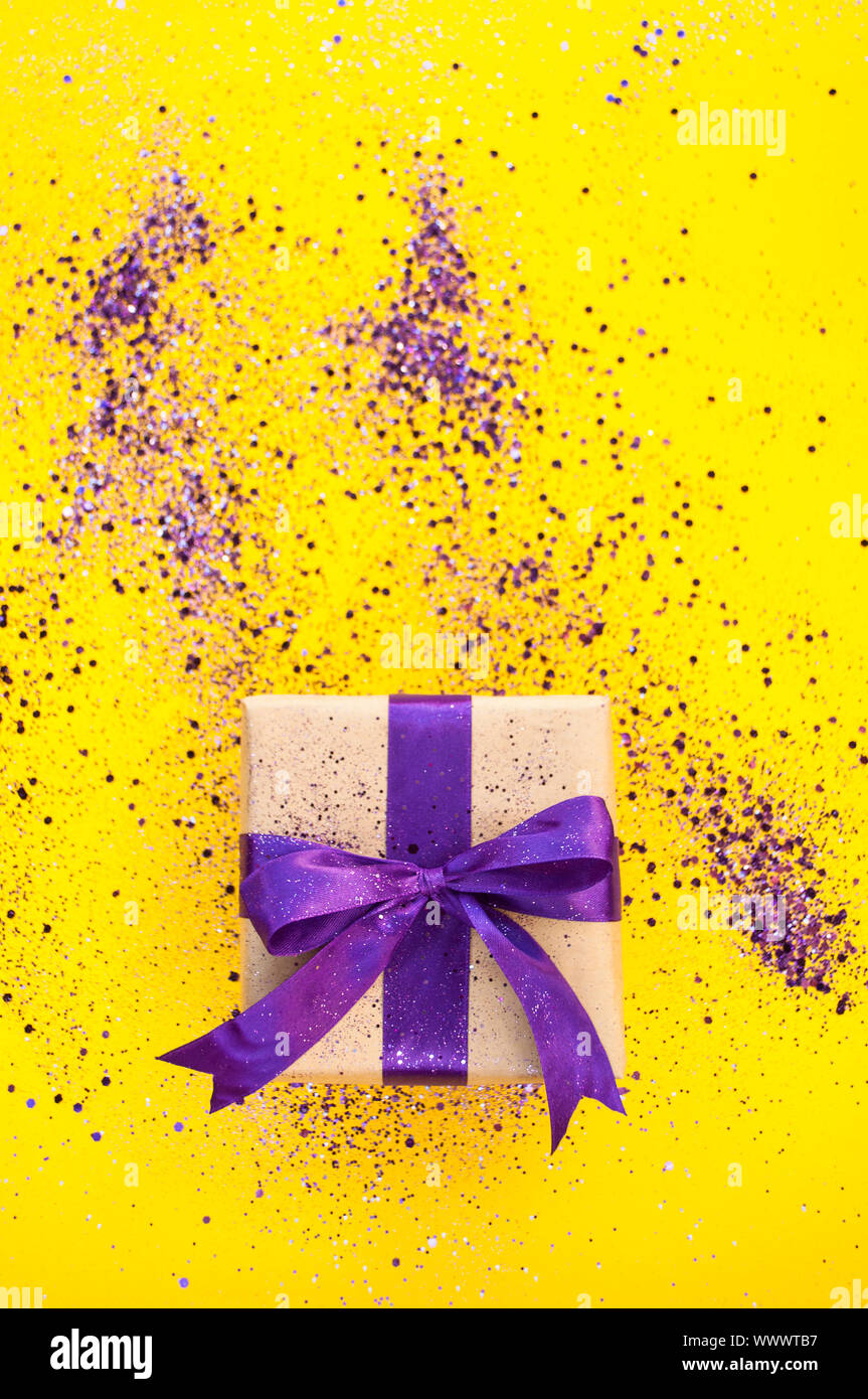 Giftbox tied with purple color ribbon on bright yellow background with glitter. Flat lay style. Stock Photo