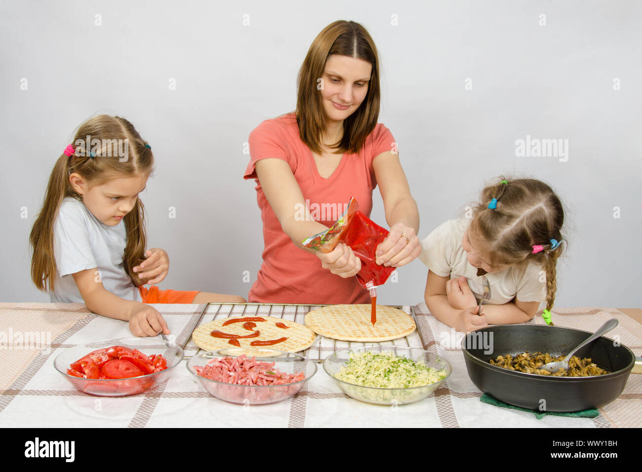 Two little girls enthusiastically watched as mum pours ketchup basis for pizza Stock Photo