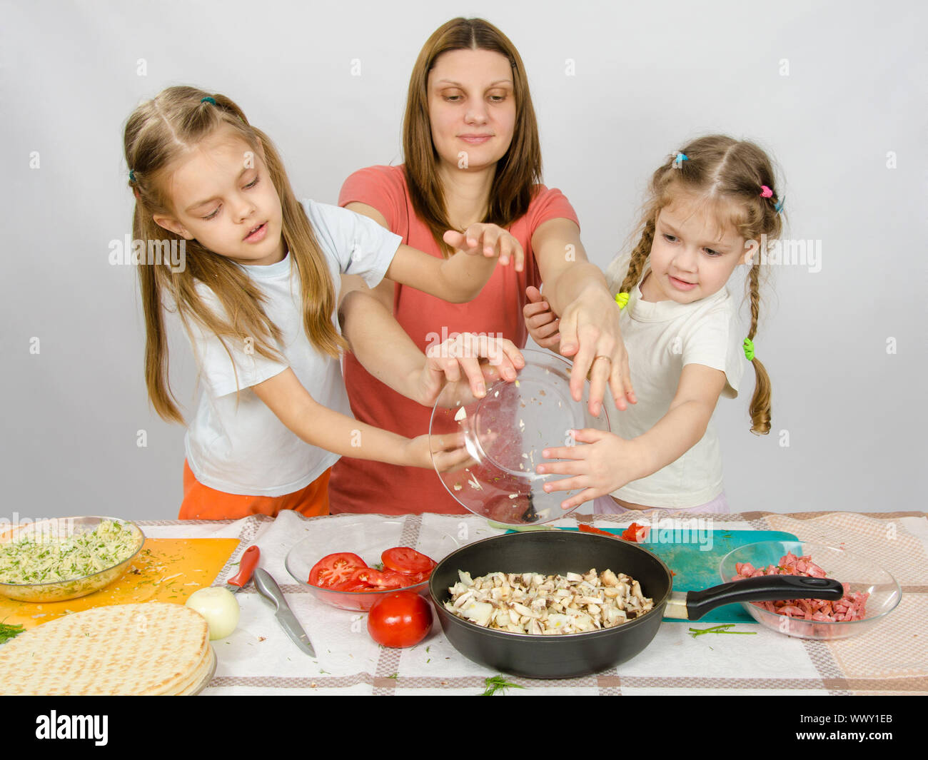 Mother with two daughters at the kitchen table with a plate of mushrooms is poured into the pan Stock Photo