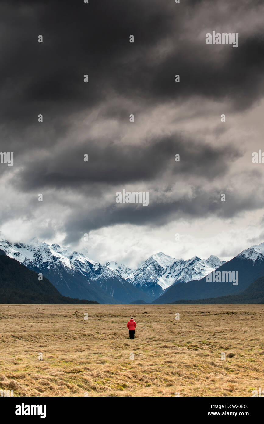Man in red coat stood looking at snow capped mountains, Fiordland National Park, UNESCO World Heritage Site, South Island, New Zealand, Pacific Stock Photo