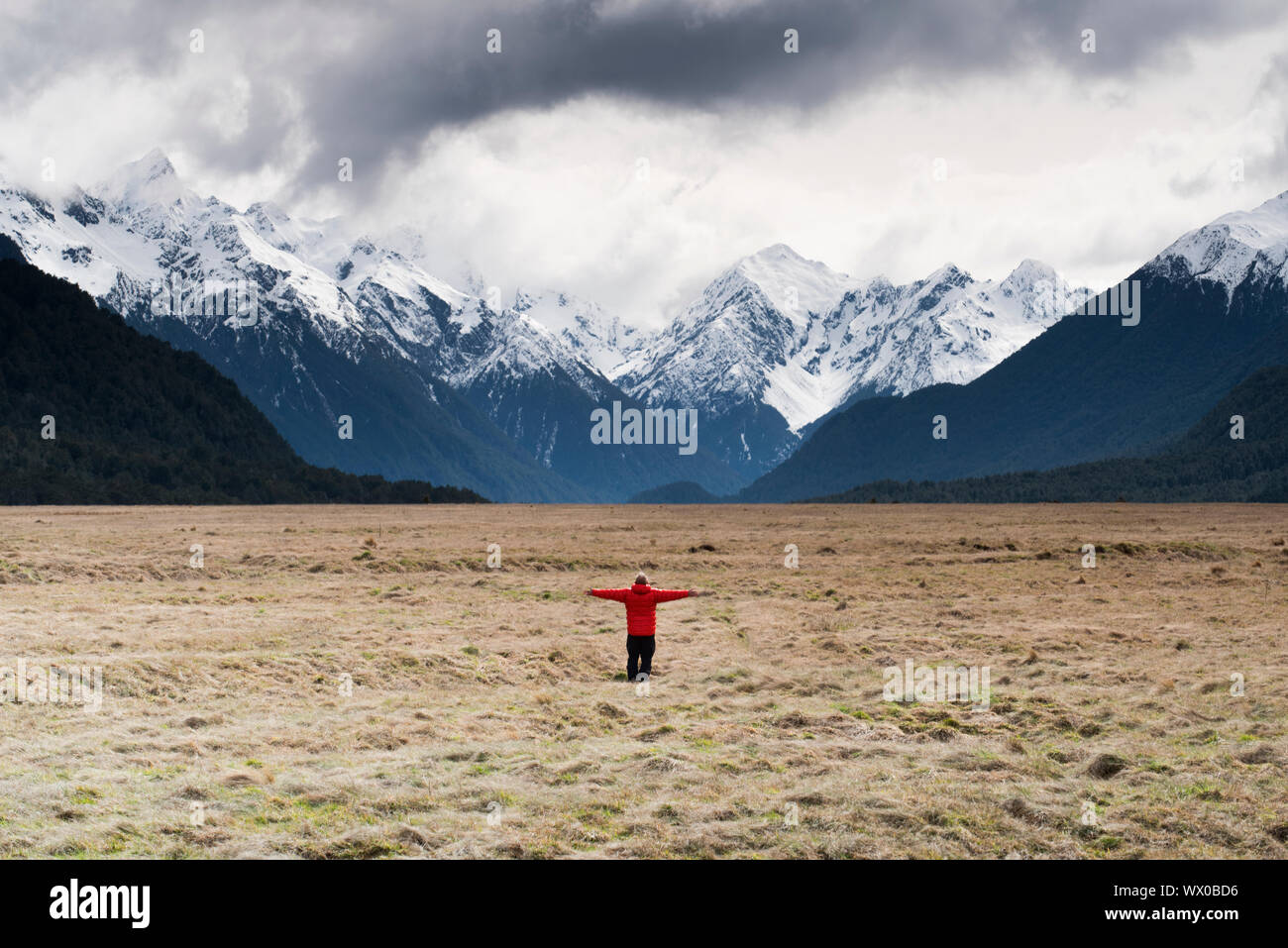 Man in red coat stood holding out his arms looking at snow capped mountains, Fiordland National Park, UNESCO, South Island, New Zealand, Pacific Stock Photo