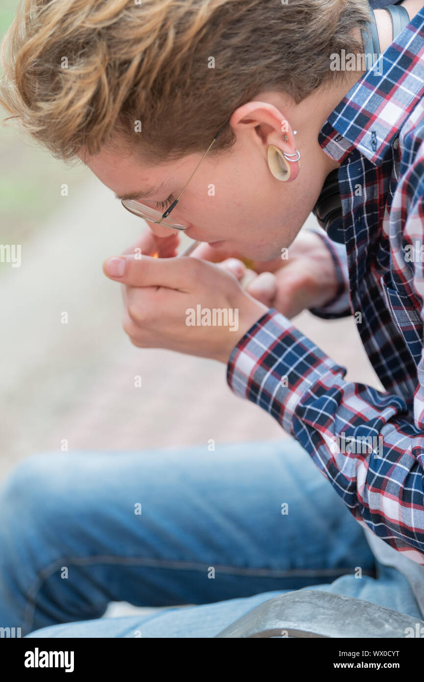 Vertical image of a relaxed blond boy wearing a square shirt lighting cigarette with the background out of focus. Stock Photo