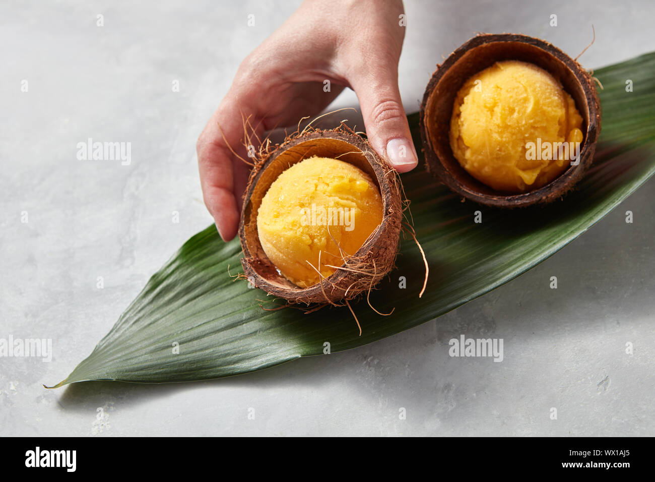 The girl's hand takes a coconut shell with mango ice cream on a gray beton background. Top view Stock Photo