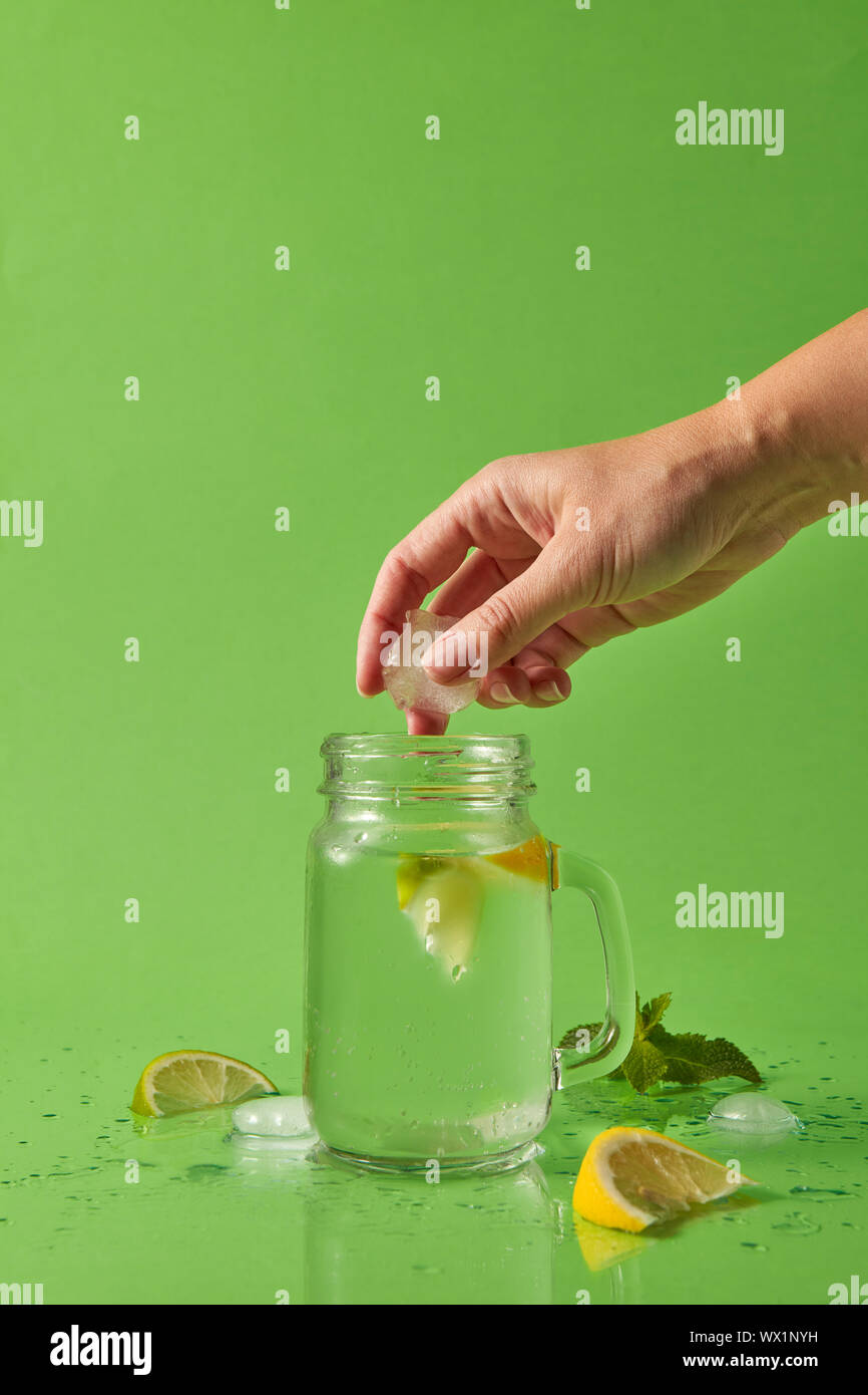 A girl's hand throws a piece of ice into a glass jar with a natural lemonade. Splash and drops on green table. Slices of lemon, Stock Photo