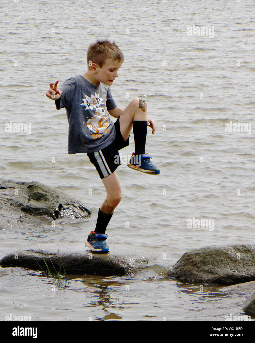 A young boy (7 yrs old) stood on one leg looking at mud on his knee Stock Photo