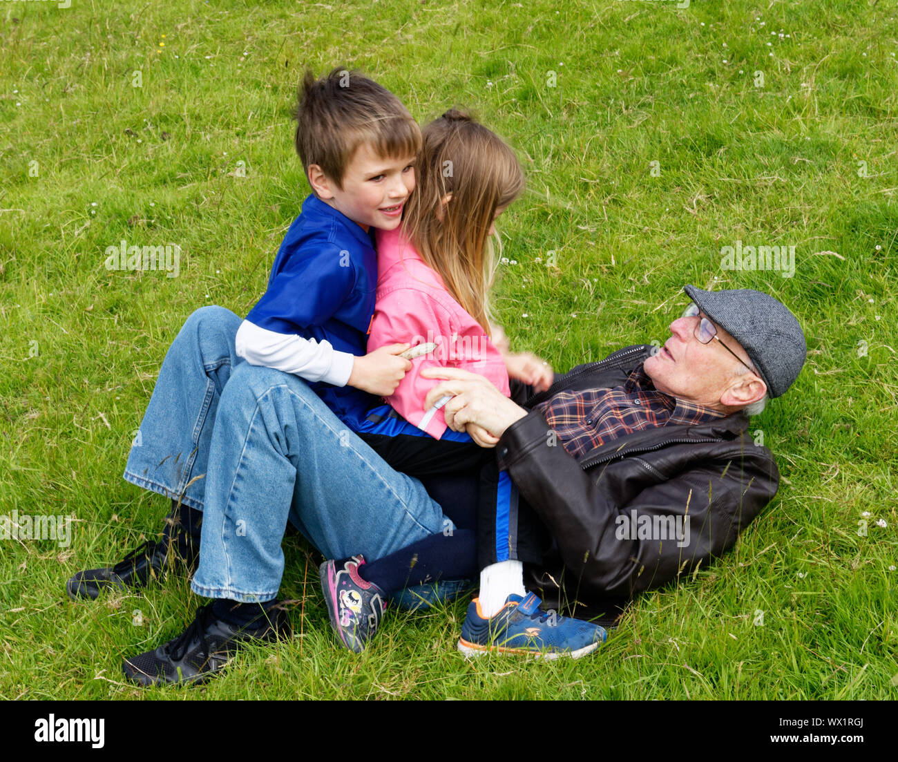 Two children (brother and sister - 7 and 5 yrs old) sat on their grandfather lying on the grass Stock Photo