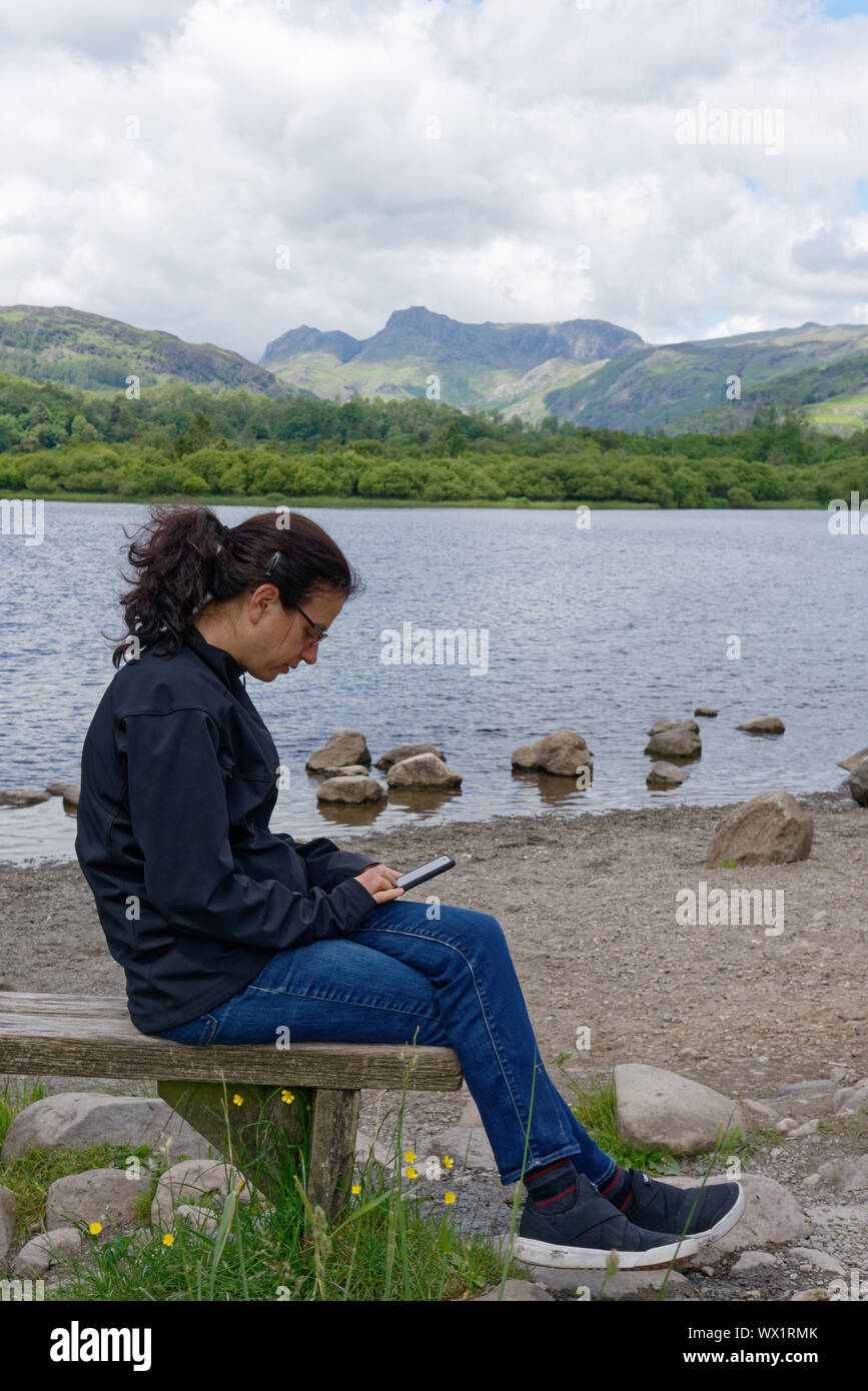 A young woman ignoring the Lake District scenery to concentrate on her cell phone, Elterwater, UK Stock Photo