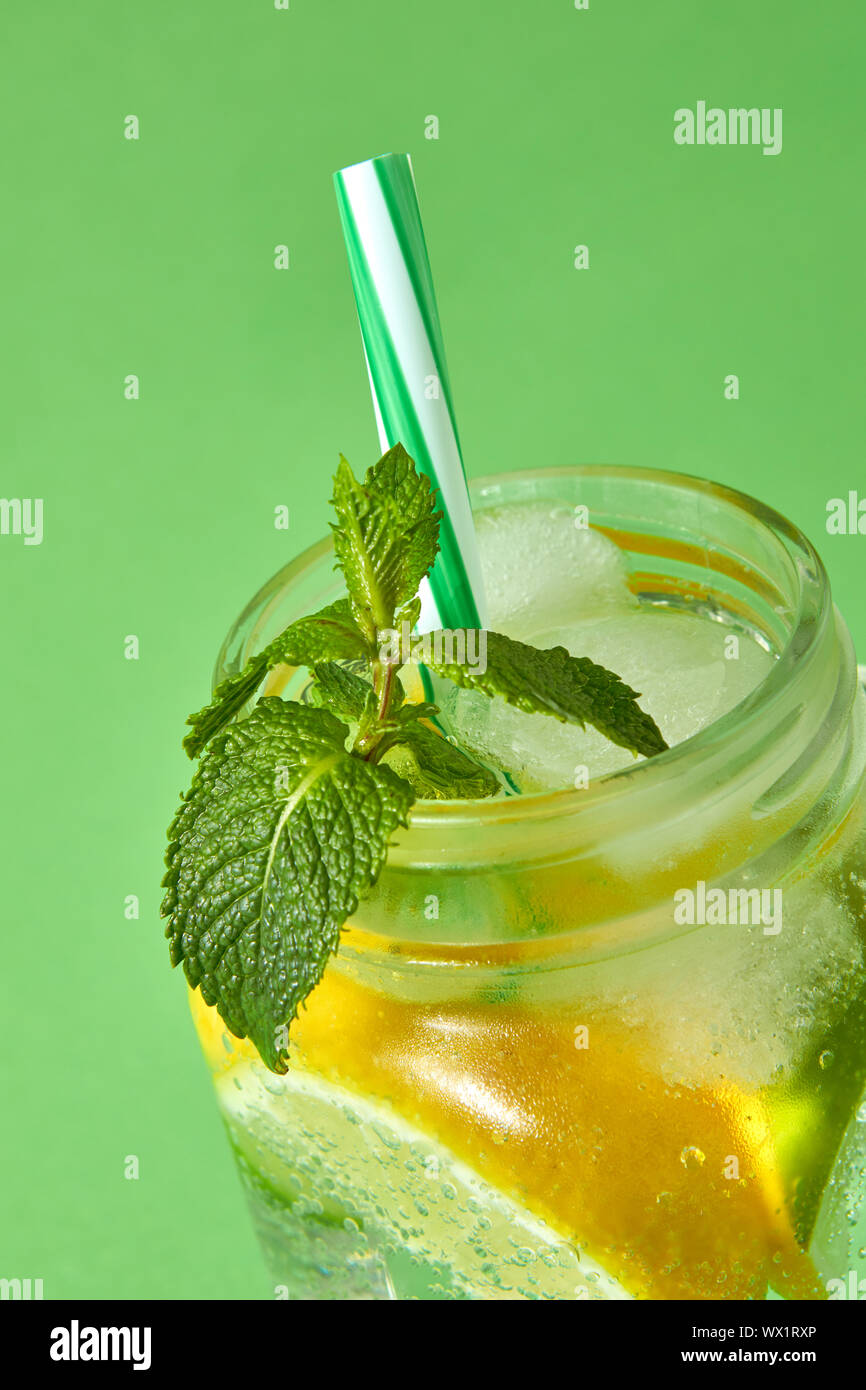 A sprig of green mint in focus in a glass jar with a cold natural homemade lemonade isolated on green. Citrus fruits slices of l Stock Photo