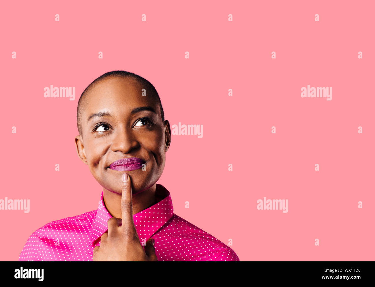 Portrait of a young woman in pink shirt with finger on mouth looking up thinking, isolated on pink studio background Stock Photo