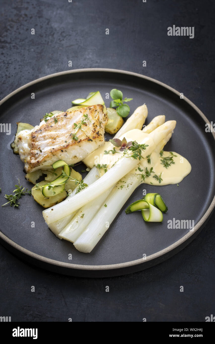 Modern German fried cod fish filet with white asparagus in hollandaise sauce with roast potatoes and sliced zucchini as top view Stock Photo