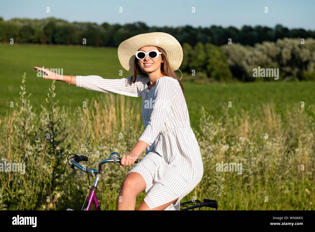 Smiling cycling girl in sunglasses and straw hat posing pointing aside with her hand Stock Photo
