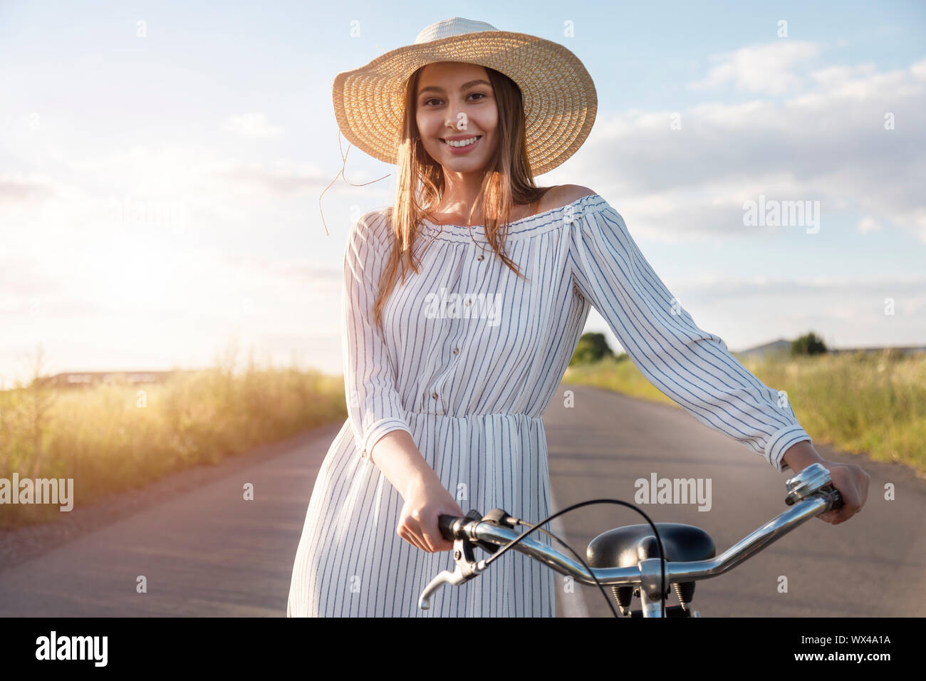 Portrait of a happy cyclist girl in white dress and straw hat on the countryside road Stock Photo