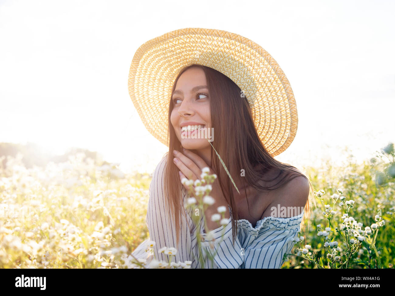 Charming brown-haired girl in straw hat and white dress enjoys sitting on the flowery meadow Stock Photo
