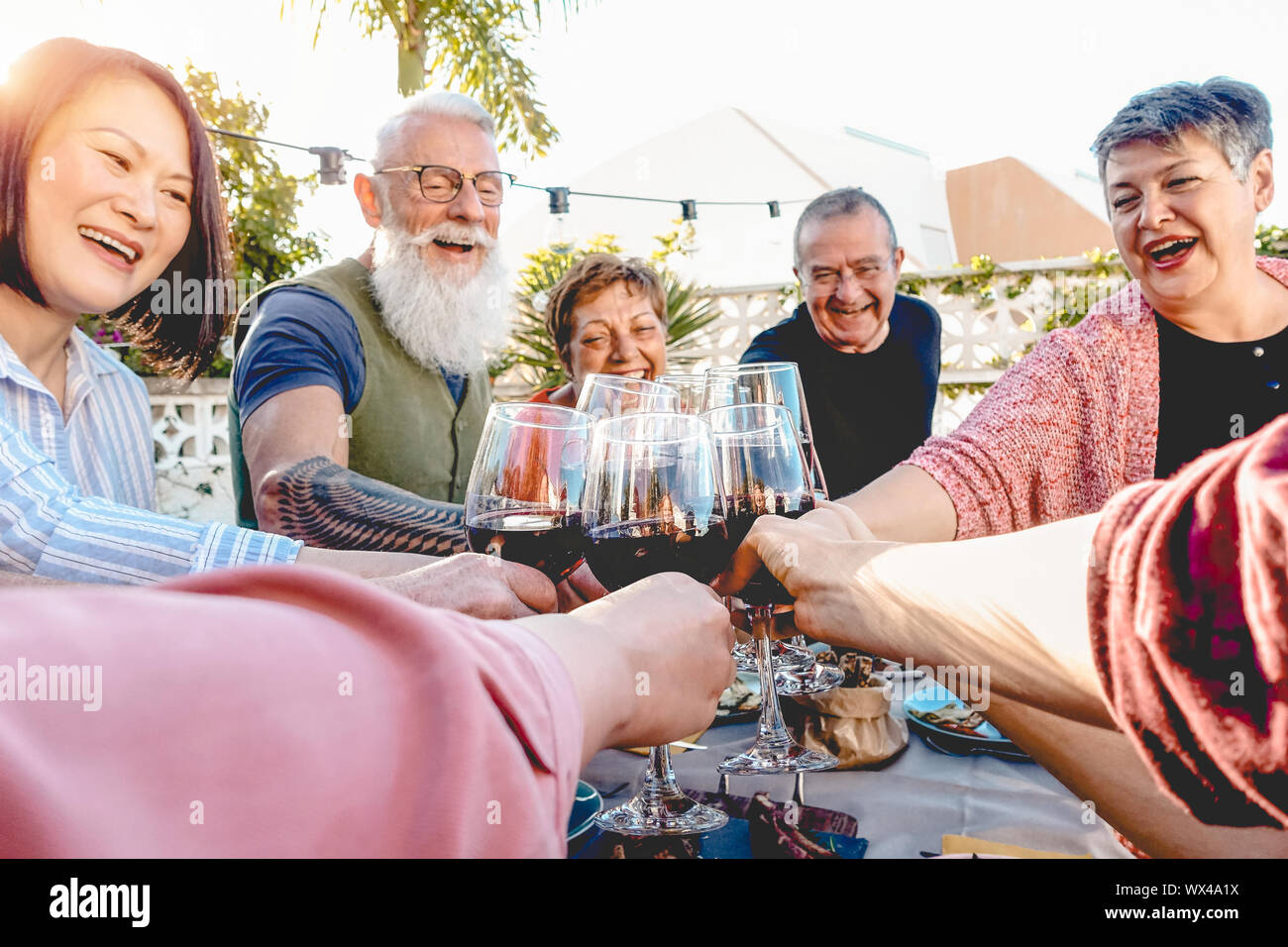 Happy senior friends toasting with red wine glasses at dinner on patio - Mature people having fun dining together outside Stock Photo