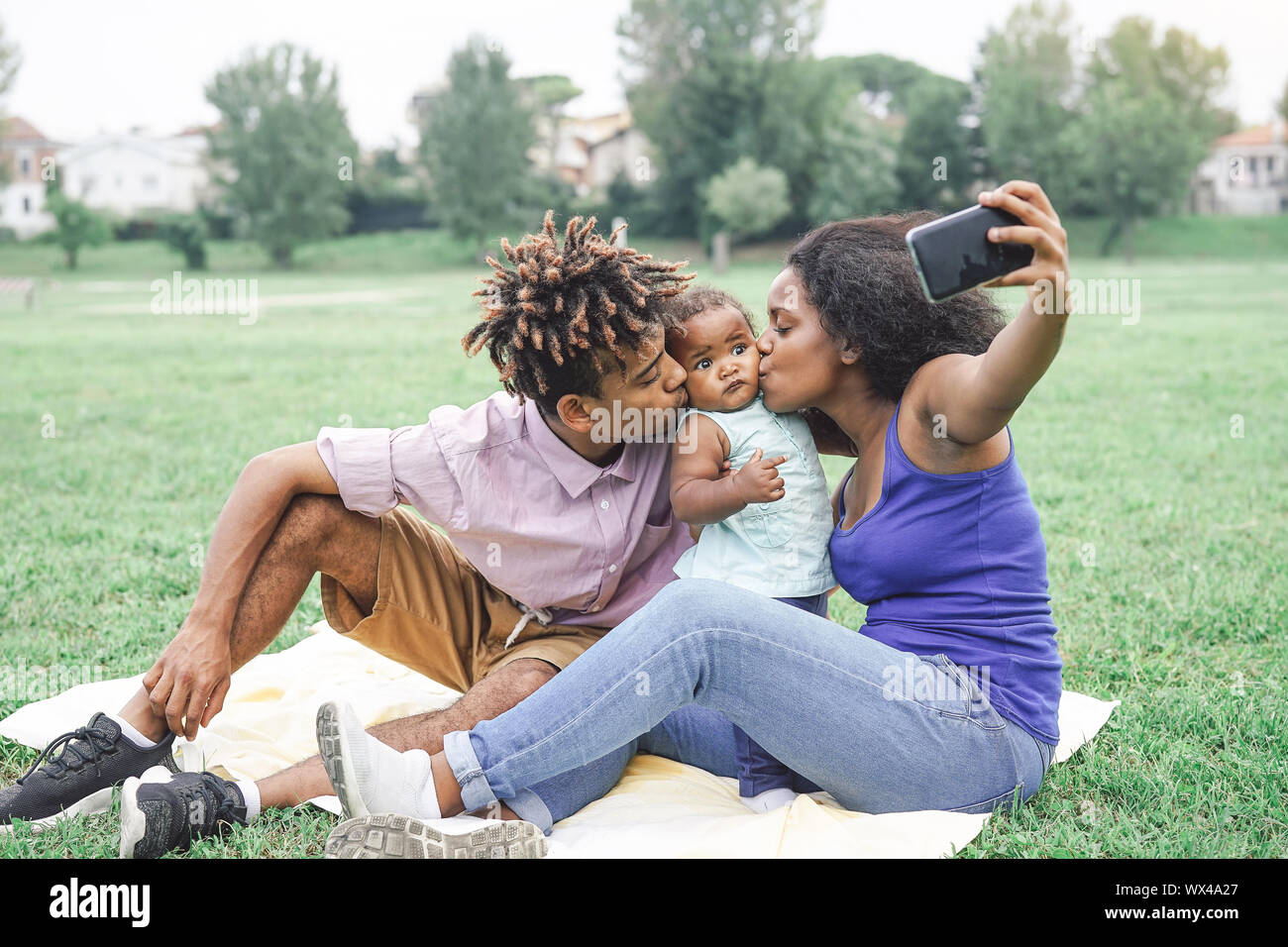 Happy african family taking a selfie with mobile smart phone camera in a public park outdoor - Mother and father having fun with their daughter Stock Photo