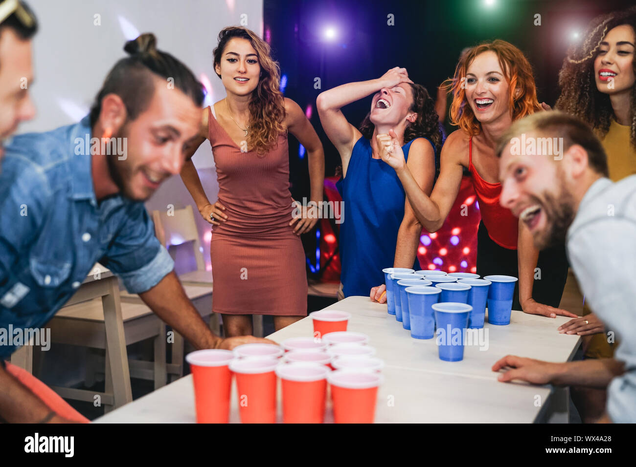 Happy friends playing beer pong in a cocktail bar - Young millennial people having fun doing party alcohol games at night pub Stock Photo