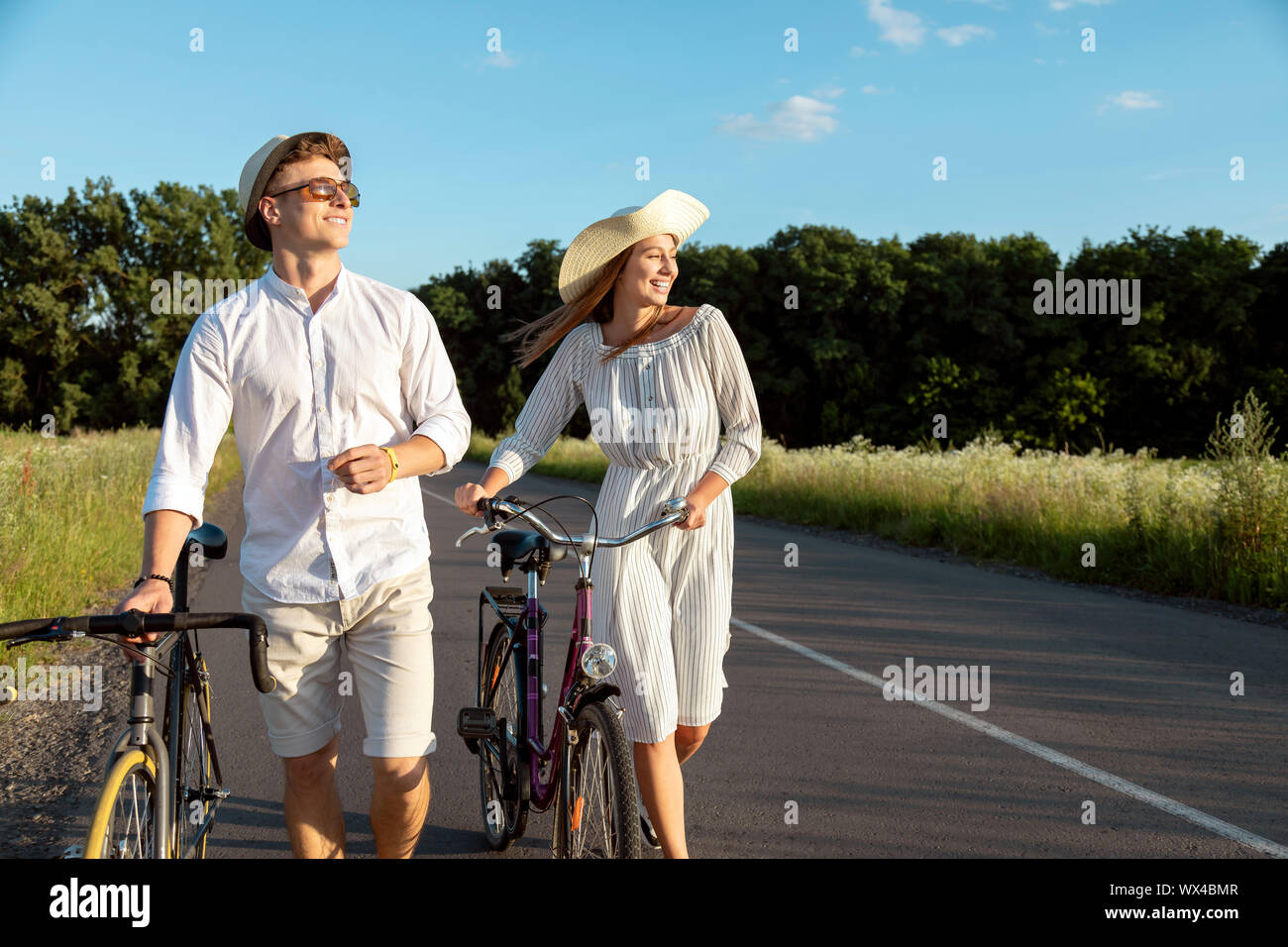 Young couple in white wheeling their bicycles along a sunny summer rural road Stock Photo