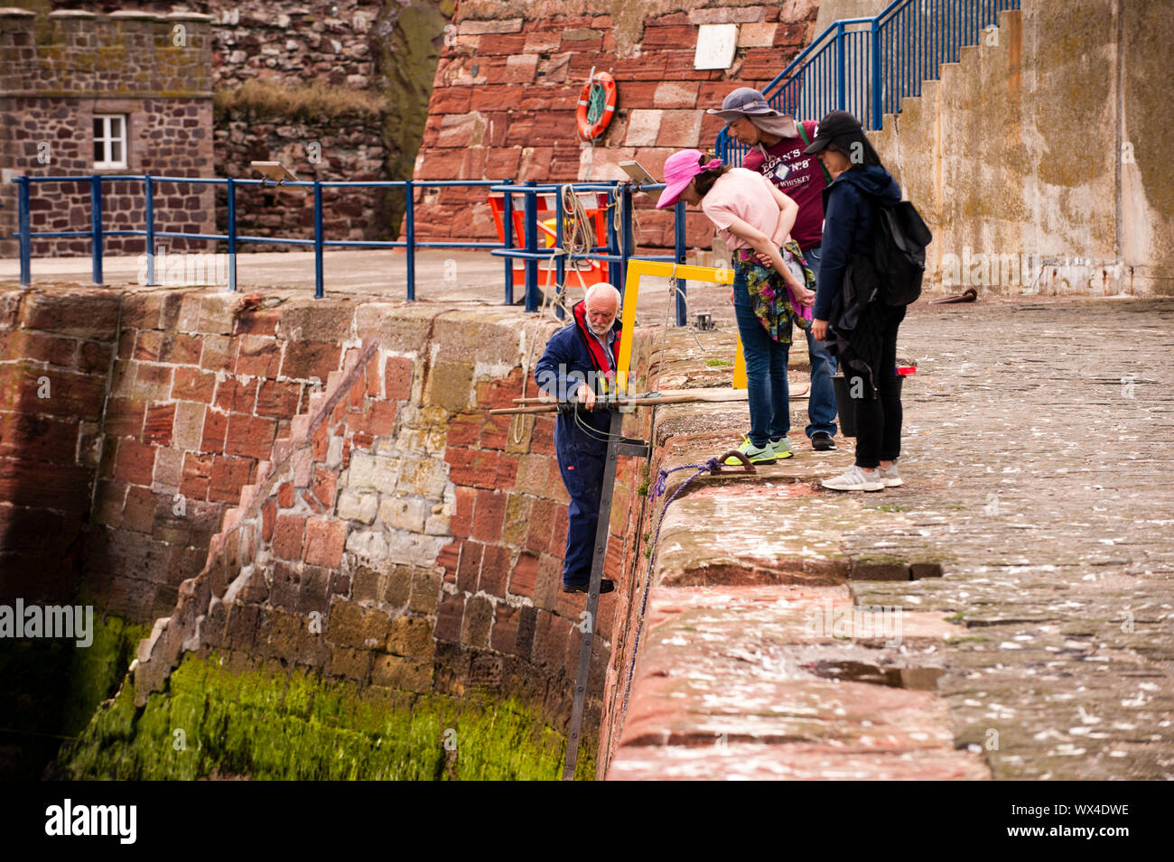 A group of tourists observe an old fisherman getting to his boat. Dunbar is a town located in the south-east of Scotland. Stock Photo