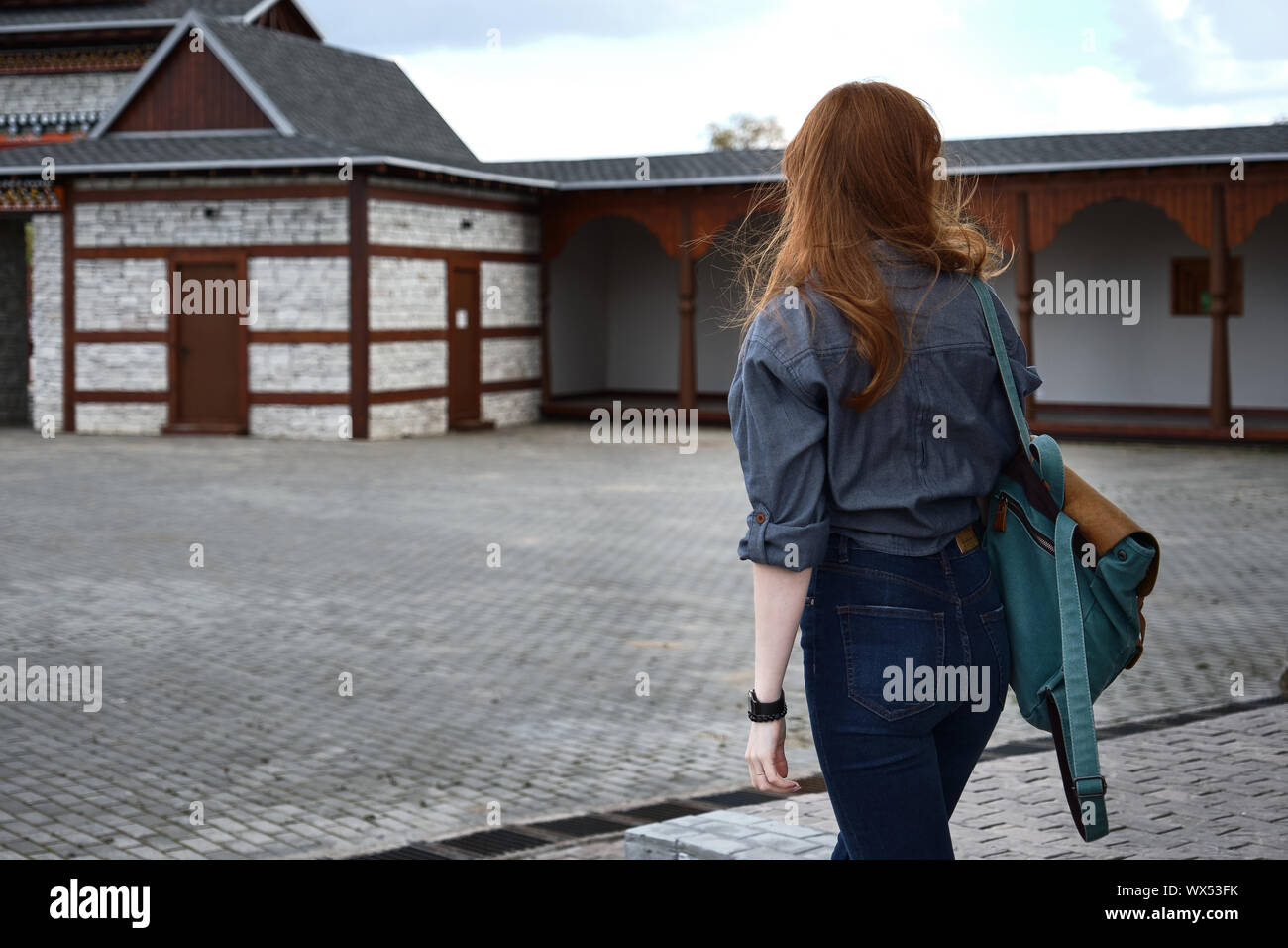 Red-haired girl in a denim shirt with a backpack is walking along an old street with stone buildings, frame from the back. Stock Photo