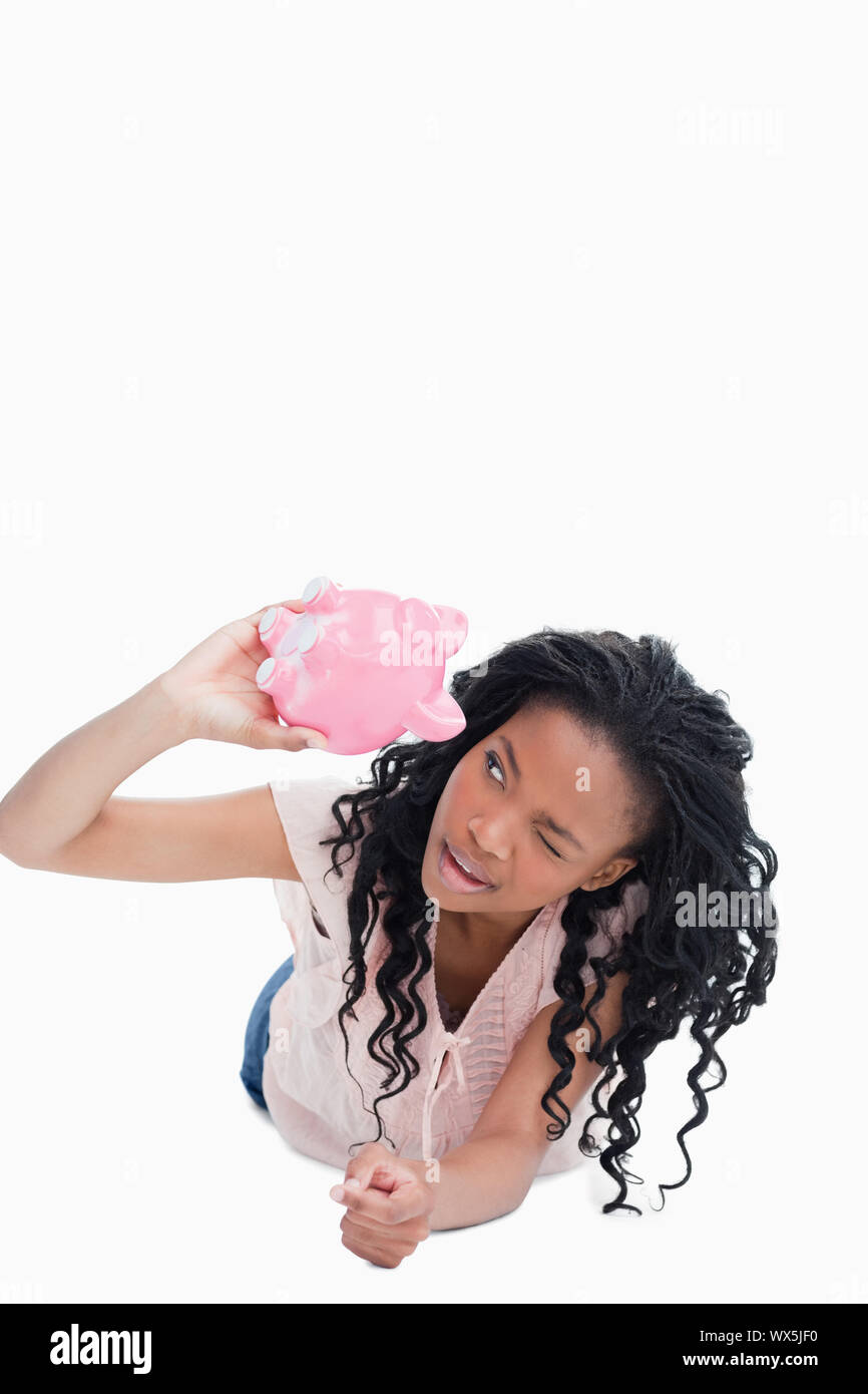 A smiling young woman is lying on the ground looking inside a piggy bank against a white background Stock Photo