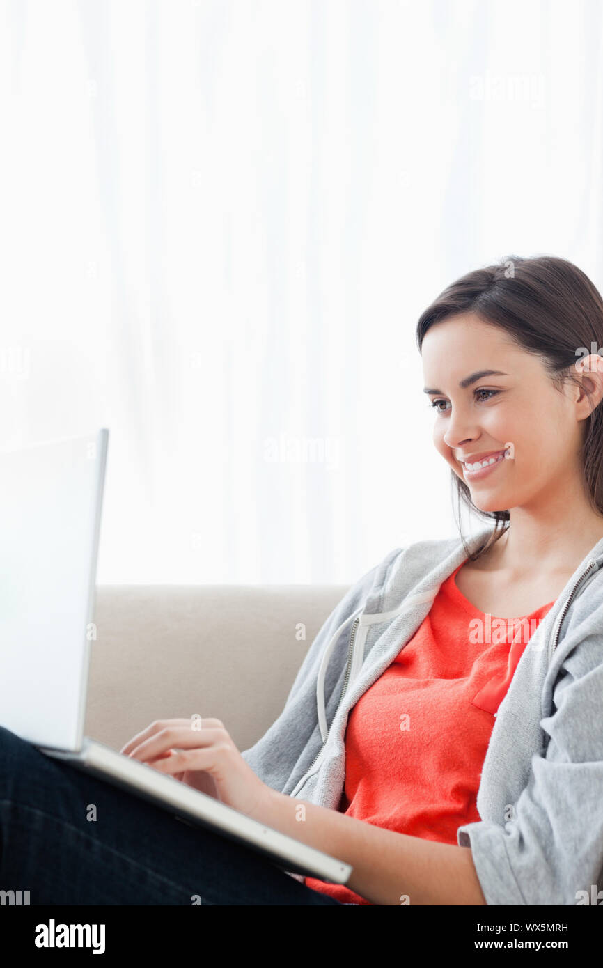 A smiling beautiful woman uses her laptop while lying on the couch ...