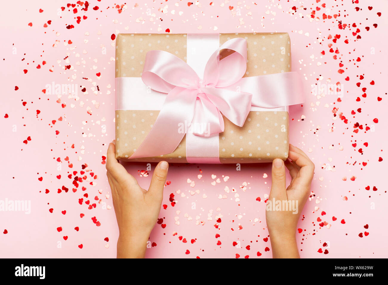 Girl's hands holding gift in kraft box with lush ribbon. Pink colors. Present for St. Valentine's day, weddings, engagements, Mother's Day, birthday, New Year, Christmas, holidays. Flat lay Stock Photo