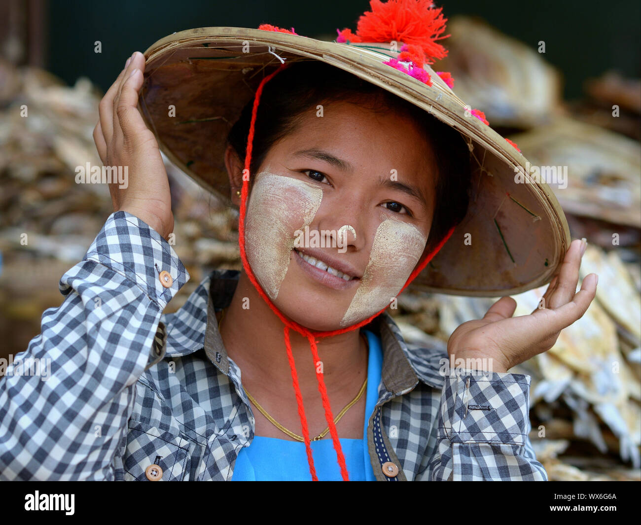 Young Burmese dry-fish saleswoman with patches of traditional thanaka face cosmetic on her nose and cheeks holds the brim of her Asian conical hat. Stock Photo