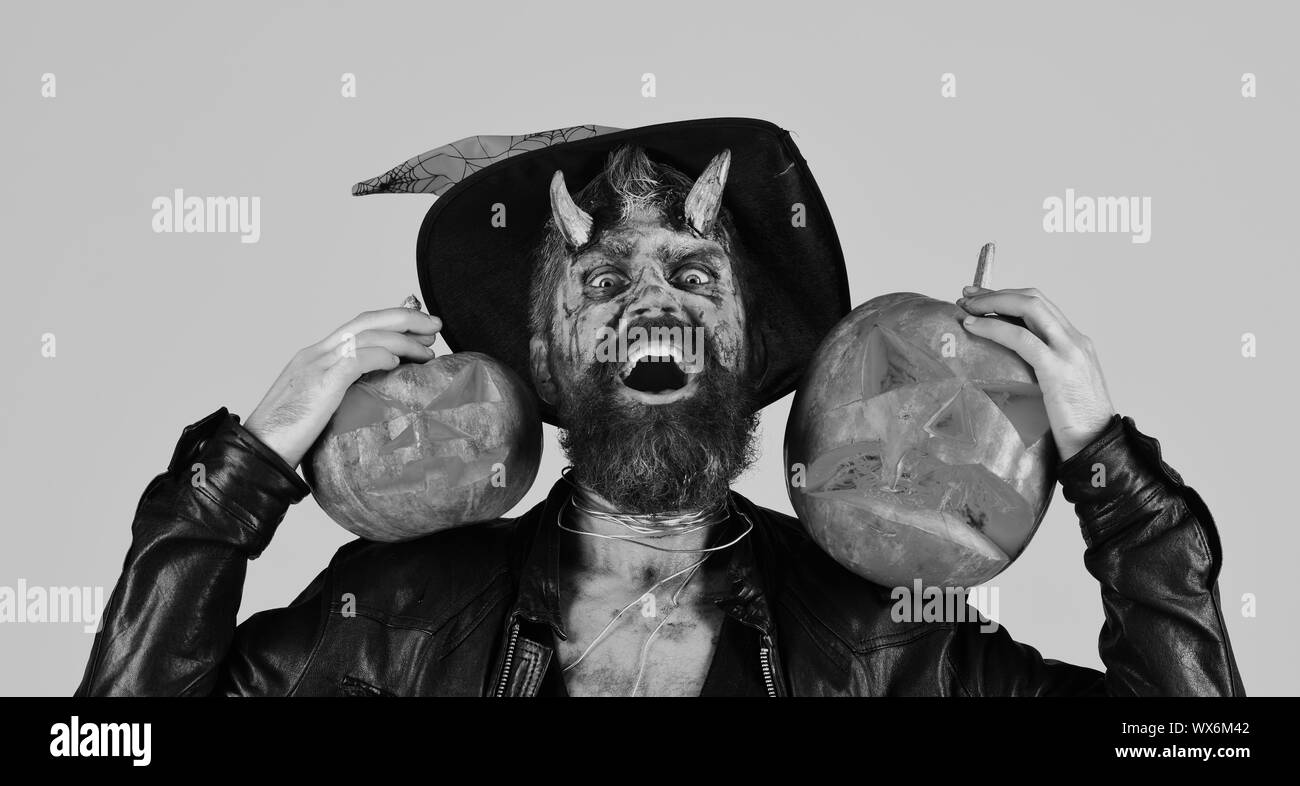 Halloween spooky party concept. Man wearing scary makeup and witchers hat  holds pumpkins on pink background.