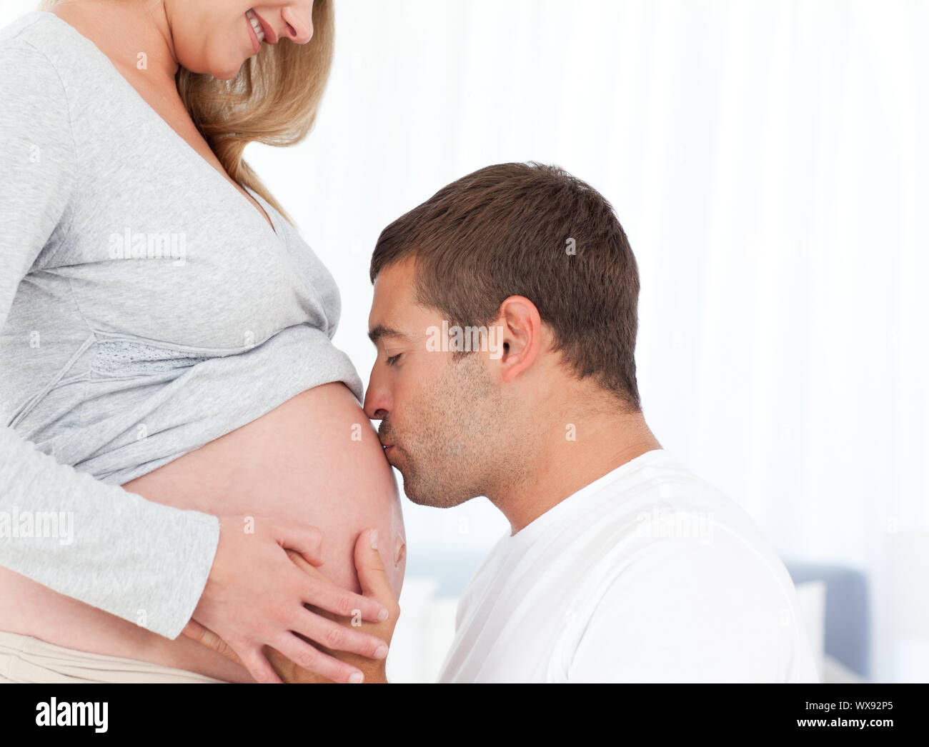 Lovely Future Dad Kissing The Belly Of His Wife Standing In The Bedroom