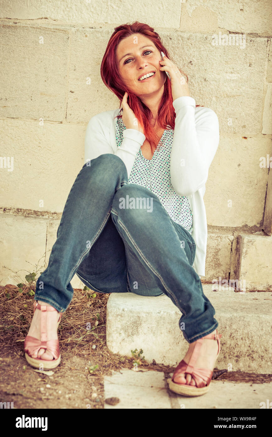 Pretty smiling redheaded woman against an old wall making a call with her cell phone Stock Photo