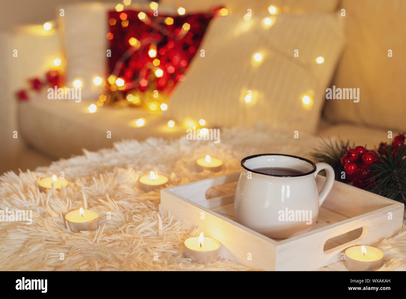 Hot cup of tea in a white mug, candles and fairy lights on a couch with pillows, modern interior Stock Photo