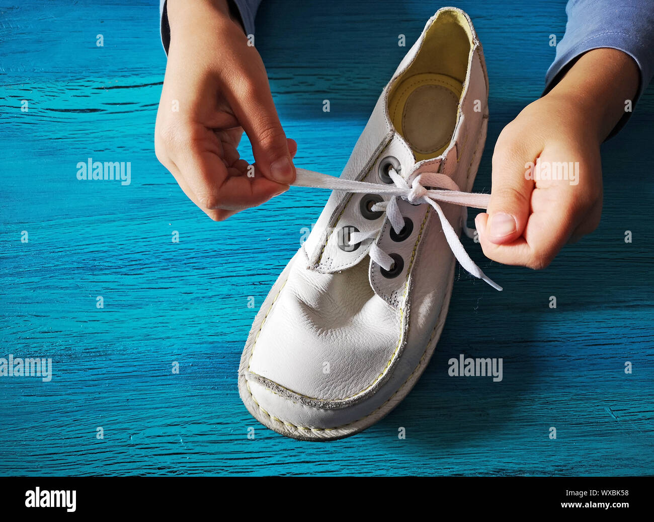 Boy is learning how to tie shoelaces, close-up on the hands and white leather shoe on turquoise wood Stock Photo