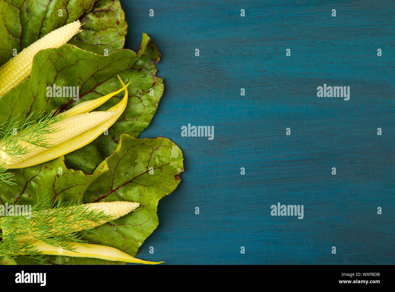 CORN IN BEET LEAF ON A WOODEN BOARD. YOUNG YELLOW EARS OF CORN IN GREEN LEAF BEETS ARE DARK BLUE WOODEN BACKGROUND. ORGANIC CERE Stock Photo