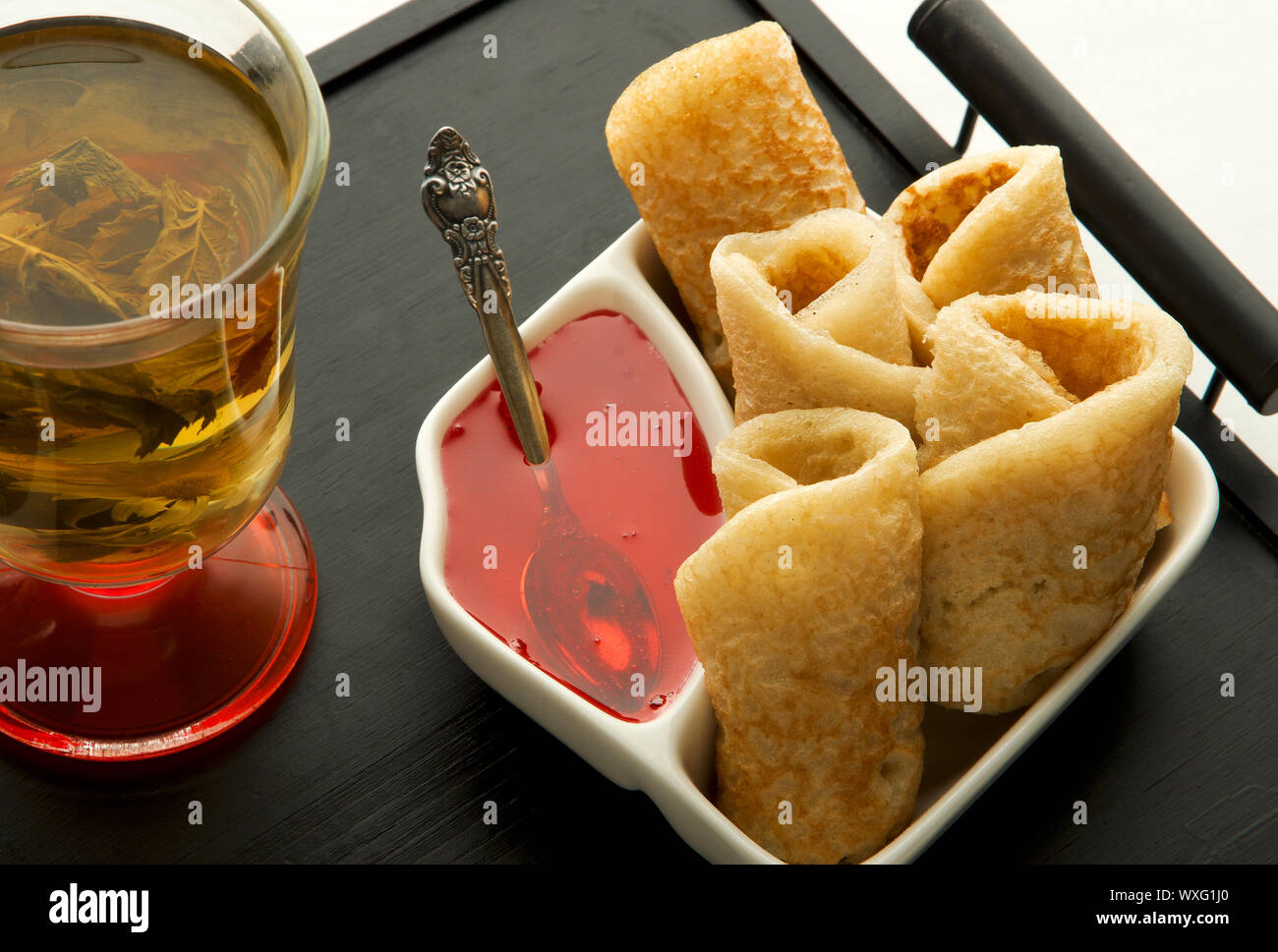 Pancakes with syrup and tea on a dark tray. Lush rosy pancakes with bright syrup on a white plate and an old spoon next to herba Stock Photo